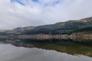 Sakin ve sakin bir dağ gölü, yemyeşil ormanları ve bulutlu bir gökyüzünün altında yuvarlanan tepeleri yansıtır. Suyun durgunluğu, manzaranın doğal güzelliğini ve huzurunu arttıran mükemmel bir ayna görüntüsü yaratır..