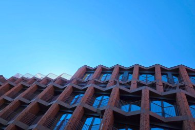 Geometrik desenler, berrak mavi gökyüzüne karşı kurulmuş çağdaş tuğla bir bina üzerinde göz alıcı bir cephe oluşturur. Açılar ve dokuların kombinasyonu modern mimari eğilimleri vurgular, kentsel tasarım estetiğini vurgular.