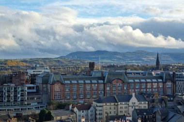 Edinburgh, İskoçya, 29 Mart 2025: Arkaplanı kayan tepeler ve dramatik bulutlu gökyüzü olan önemli bir mimari binanın yer aldığı tarihi şehir manzarası. Kentsel gelişim ve doğal güzelliğin yan yana getirilmesi kültürel mirası vurguluyor.