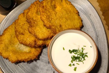 Seramik tabakta taze otlarla kaplı bir kase kremalı ekşi kremanın yanında altın rengi çıtır patatesli latke servis ediliyor. Bu latkeler geleneksel ve lezzetli mutfağın vücut bulmuş halidir, şenlikli toplantılar veya aile yemekleri için idealdir..
