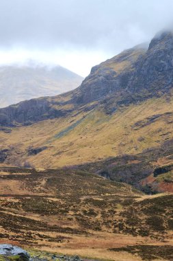 Görkemli dağ manzarası bulutlu gökyüzünün altında engebeli tepeler sergiliyor. Manzara kayalık arazi, çimenli yamaçlar ve bastırılmış renk paleti karışımı içerir, doğa ve seyahat temaları için mükemmeldir..
