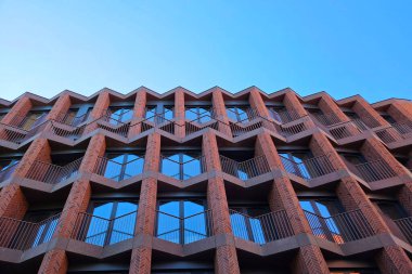 Modern bina cephesinde karmaşık geometrik desenler ve kırmızı tuğla ve cam elementlerle simetri gösteriliyor. Perspektif, çağdaş kentsel mimarinin vurgulandığı açık mavi gökyüzüne karşı tekrarlanan üçgen motifleri vurgular.