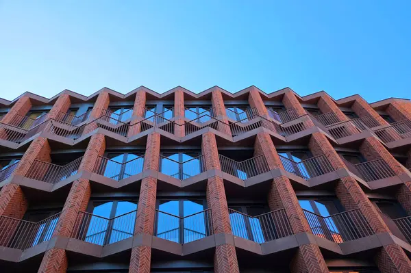 Modern bina cephesinde karmaşık geometrik desenler ve kırmızı tuğla ve cam elementlerle simetri gösteriliyor. Perspektif, çağdaş kentsel mimarinin vurgulandığı açık mavi gökyüzüne karşı tekrarlanan üçgen motifleri vurgular.