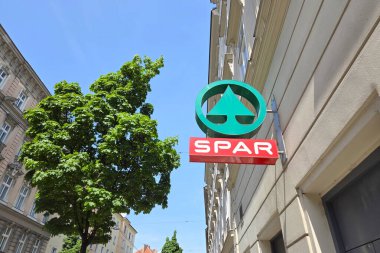 Viyana, Avusturya, 3 Mayıs 2025: Açık mavi gökyüzünün altındaki bir şehir binasının ön cephesine spor süpermarket tabelası asıldı. Canlı yeşil ağaç, mimari ortama, somutlaşmış şehir yaşamına ve ticari kolaylığa doğal bir zıtlık ekler..