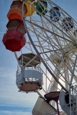Parlak mavi gökyüzüne karşı dizilmiş renkli gondolları olan canlı bir dönme dolabın görüntüsü. Yapı canlı renklerin bir karışımını sergiliyor, yaz fuarları ve lunaparklara özgü neşeli ve canlı bir atmosfer yaratıyor..