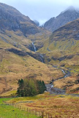 İskoçya dağlarındaki engebeli bir dağdan çağlayan şelalenin sakin bir manzarası, yemyeşil ve kayalık arazilerle çevrili. Bulutlu gökyüzü manzaraya dramatik bir dokunuş ekler..