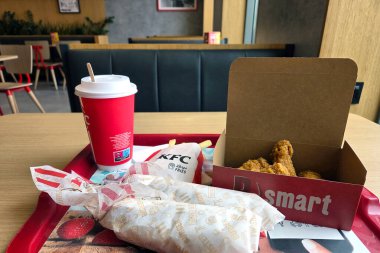 Wroclaw, Polonya, 12 Nisan 2025: Çağdaş bir restoran ortamında kızarmış tavuk, paket, patates kızartması ve soda bardağının yer aldığı iyi düzenlenmiş bir fast food yemeği. Sahne, hareketli ve davetkar bir atmosferde rahat ve rahat bir akşam yemeğini ön plana çıkarıyor..