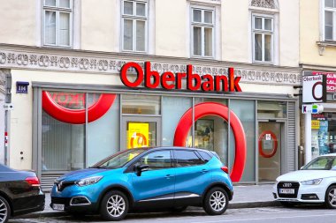 Viyana, Avusturya, 4 Mayıs 2025: Canlı bir şehir sahnesinde belirgin kırmızı logolu bir banka cephesi yer alıyor. Aralarında mavi bir cip ve gümüş bir sedan 'ın da bulunduğu iki park edilmiş araba, Avrupa' da bir caddede günlük şehir yaşamını örnek alarak önde duruyorlar..