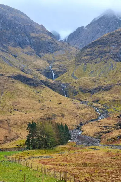 İskoçya dağlarındaki engebeli bir dağdan çağlayan şelalenin sakin bir manzarası, yemyeşil ve kayalık arazilerle çevrili. Bulutlu gökyüzü manzaraya dramatik bir dokunuş ekler..