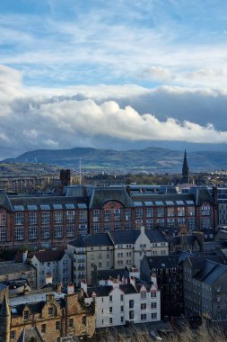 Edinburgh, İskoçya, 29 Mart 2025: Şehrin tarihi mimarisini çeşitli çatı stilleriyle gösteren panoramik bir manzara. Arka planda, uzak dağlar canlı, bulutlarla dolu bir gökyüzü altında yükseliyor, kentsel ve doğal elementleri harmanlıyor..