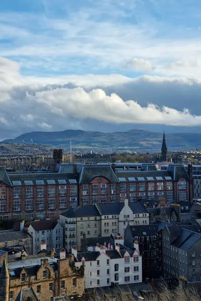 Edinburgh, İskoçya, 29 Mart 2025: Şehrin tarihi mimarisini çeşitli çatı stilleriyle gösteren panoramik bir manzara. Arka planda, uzak dağlar canlı, bulutlarla dolu bir gökyüzü altında yükseliyor, kentsel ve doğal elementleri harmanlıyor..