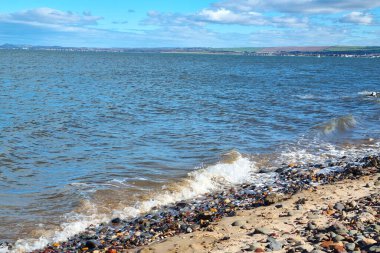 Kayalık kıyı şeridi, yumuşak dalgalar çakıl taşlarına çarpıyor, geniş ve parlak bir gökyüzünün altında. Uzak tepeler ve geniş deniz doğa temaları için ideal sakin bir kıyı manzarası yaratır..