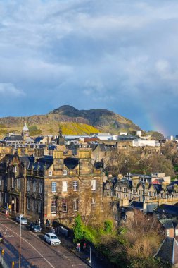 Edinburgh, İskoçya, 29 Mart 2025 tarihi taş binalar ve uzak bir şatonun yer aldığı Edinburgh manzarası. Bereketli tepeler, bulutlu bir gökkuşağından yükselen bir gökkuşağı tarafından vurgulanan bir zemin görevi görür. Kentsel sokaklar coşkuya karakter katıyor 