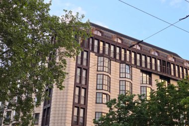 Viyana, Avusturya, 3 Mayıs 2025: Modern yüksek binalar çağdaş tasarım ve yemyeşil alanları harmanlıyor. Yapı, cam ve tuğla elementlerin bir karışımı ile benzersiz bir cephe içerir, doğal ışık ile aydınlatılmış, berrak mavi bir gökyüzü altında,