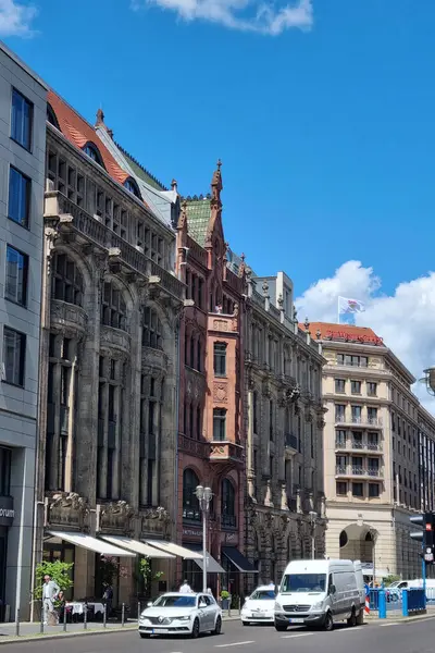 Berlin, Almanya, 26 Mayıs 2025: Canlı bir şehir sokak sahnesi parlak mavi gökyüzü altında tarihi mimari yapılar sergiliyor. Beyaz araçlar yol boyunca giderken, insanlar bir kaldırım kafesinde yemek yerken görülüyor ve şehre yaşam ve enerji katıyor.