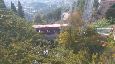 Kırmızı bir teleferik yoğun ormanlar ve tepe yapılarıyla çevrili yeşil dağlık bir alanda seyahat eder. Arkaplan, berrak mavi bir gökyüzü altında, sükunet ve macerayı yansıtan geniş bir şehir manzarasını içeriyor..