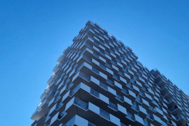 Bulutsuz mavi gökyüzüne karşı geometrik desenler gösteren modern bir gökdelenin soyut cephesi. tasarım, kentsel yapıdaki yenilikleri yansıtan, eşsiz kareli görünümlü çağdaş mimariyi vurgular..