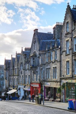 Edinburgh, İskoçya, 30 Mart 2025: Geleneksel binalarla birlikte tarihi mimarinin yer aldığı Avrupa sokak sahnesi. Parlak, bulutlu bir gökyüzünün altında gezinen insanlar, parke taşı caddesini çevreleyen canlı vitrinler,