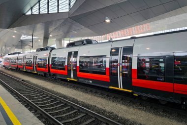 Viyana, Avusturya, 5 Mayıs 2025: Pistte hareketli bir şehir istasyonunda parlak kırmızı ve gri bir tren duruyor. Genel mimari açısal çizgiler ve geniş pencerelerle modern olup, toplu taşıma merkezlerindeki çağdaş tasarım eğilimlerini yansıtmaktadır..