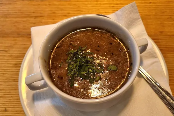 Taze otlarla kaplı, dumanı tüten bir mantar çorbasının yakın plan görüntüsü. Beyaz seramik bir fincanda sunuluyor. Peçeteye konmuş, ahşap bir masa arkaplanı var. Sıcak bir yemek atmosferi yaratıyor..