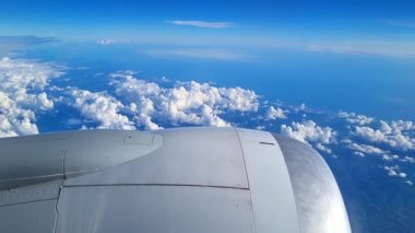 Bir uçak penceresinden yüksek irtifa görüntüsü canlı mavi gökyüzüne karşı geniş, tüylü, beyaz bulutlar sergiliyor. Uçak kanadı görünür, uçuş ve seyahat deneyimini vurguluyor..