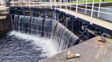 Metal bir geçitle çevrili endüstriyel bir barajın üzerinde çağlayan sular. Sahne doğal ve endüstriyel elementleri harmanlıyor, titizlikle tasarlanmış yapı üzerinde suyun akışını gösteriyor, doğa ve teknoloji arasındaki zıtlıkları gösteriyor..