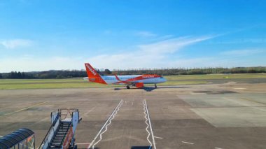 Edinburgh, İskoçya, 1 Nisan 2025: Özel turuncu ve beyaz üniformalı ticari bir uçak geniş bir havaalanının pistine park edilmiştir. Mavi gökyüzü ve çevreleyen yeşillik sahneleri güzelleştiriyor, seyahat temaları için ideal..