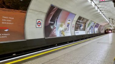 Londra, İngiltere, 18 Mayıs 2025: Uzaktan yaklaşan bir trenin yer aldığı bir Londra yeraltı platformu. Eğri duvarlarda seyahat ve teknoloji temaları da dahil olmak üzere çeşitli reklamlar sergileniyor.. 