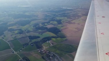 Uçsuz bucaksız tarlalar, ormanlar ve bir uçak penceresinden görüldüğü gibi temiz bir gökyüzünün altında dolambaçlı bir nehir sergiliyor. Uçağın kanadı kısmen görülebilir olup, uçuş ve keşif hissini arttırır..