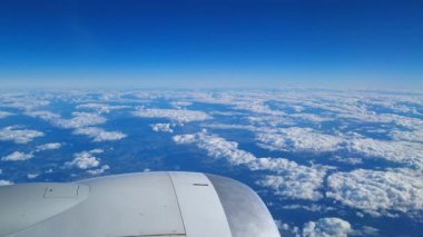 Manzaralı bir hava manzarası, kabarık bulutların ve açık mavi gökyüzünün arka planına karşı bir uçak kanadı içerir. Bu yüksek irtifa perspektifi seyahat, macera ve yeryüzünün üzerinde uçmanın huzurunu taşır..