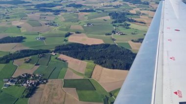 Bir uçak penceresinden, çeşitli tarım arazilerinden yeşil ve kahverengi alanların hava görüntüsü. Uçak kanadı görünür, seyahat ve keşif temasını geliştirir..