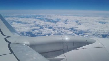 Bir uçak penceresinden, kabarık bulutların ve engin mavi gökyüzünün ardında bir kanadı göstererek, hava yolculuğunun özünü ve yükseklikte uçmanın dingin güzelliğini yakalıyor..