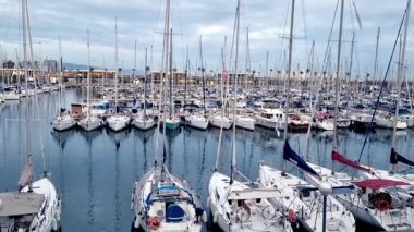 Barselona, İspanya, 3 Ocak 2025: Bulutlu bir gökyüzünün altında demirlemiş sayısız yelkenliyle dolu geniş bir marina. Sakin su damarları yansıtır, seyahat, macera ve eğlence temalı tasarımlar için mükemmel bir deniz manzarası yaratır..
