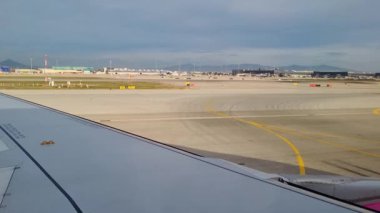 Barselona, İspanya, 4 Ocak 2025: Bir uçak penceresinden manzaralı bir manzara açık gökyüzü, uzak binalar ve ufukta dağlarla geniş bir havaalanı pistini gösteriyor. Seyahat temalı tasarım, baskı ve dijital ortam için ideal.
