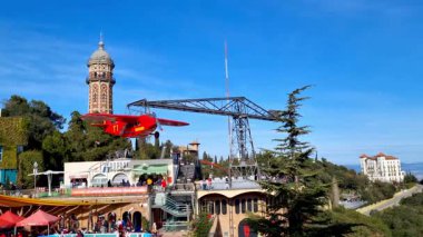 Barselona, İspanya, 2 Ocak 2025: Canlı kırmızı bir uçak yolculuğunun, arka planda tarihi bir kulenin ve hareketli bir kalabalığın yer aldığı heyecan verici bir lunapark sahnesi. Açık mavi gökyüzü, heyecan verici bir açık hava sunarak, canlı atmosferi arttırır.
