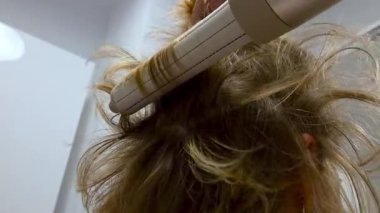 Saç maşasının yakın plan görüntüsü, sarı saçlarda kıvrımlar oluşturma sürecini gösteriyor. Saç şekillendirme tekniklerini, saç dokusunu ve profesyonel buklelerin elde edilmesinde kullanılan araçları vurgular.