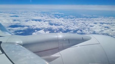 Büyüleyici bir hava manzarası, bir uçak penceresinden görüldüğü gibi, dağ zirveleriyle karışmış tüylü bulutları gösteriyor. Uçak motorları ve kanatları açık mavi gökyüzüne karşı kısmen görünür. Gezgin şehvet ve macera hissi uyandırıyor..