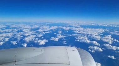 Bir uçak penceresinden yüksek irtifa görüntüsü açık mavi gökyüzünün altında geniş bir beyaz kabarık bulutlar sergiliyor. Uçak motoru kısmen görünür durumda, bu da hava yolculuğu, macera ve keşif anlamına geliyor..