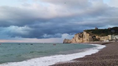 Gün batımında bulutlu bir gökyüzüne karşı dramatik uçurumların olduğu sahil sahnesi. Ön planda çakıl taşı plajı ve sakin okyanus dalgaları sakin ama çarpıcı bir manzara yaratır. Küçük tekneler suda yüzer, denizcilik cazibesi katar..