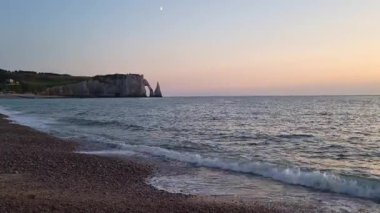 Etretat ikonik uçurumları günbatımının yumuşak renklerinde yıkanır ve huzurlu bir deniz manzarası yaratır. Kibar dalgalar çakıl taşı sahilini kaplıyor, doğal güzelliğin huzurlu bir sahnesini sunuyor, seyahat ve manzara fotoğrafçıları için mükemmel..
