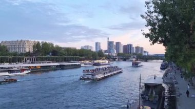 Paris, Fransa, 3 Ağustos 2025: Hareketli bir nehir sahnesinde gün batımında canlı bir şehir su yolu boyunca birden fazla tekne yer alır. Ufuk çizgisi modern gökdelenlerle benekli ve yemyeşil deniz kıyılarını çevreleyerek, harmonik bir harman oluşturuyor.