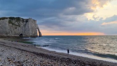 Etretat sahilinin manzarasında gün batımında dramatik bir gökyüzünün altında tebeşir kayalıkları yükselir. Altın ufuk, dokulu çakıl taşları ve dinamik okyanus dalgaları arasındaki etkileşim çarpıcı bir görsel deneyim sunuyor..