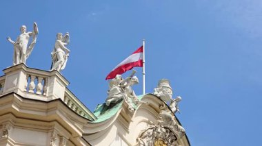 Viyana, Avusturya, 3 Mayıs 2025: Süslü tarihi bir binanın üzerinde sallanan Avusturya bayrağı klasik heykellerle süslenmiş, canlı mavi bir gökyüzüne karşı dizilmiş. Mimaride, geleneksel Avrupa tasarım unsurlarının yer aldığı karmaşık ayrıntılar yer alıyor..