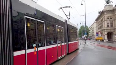 Viyana, Avusturya, 5 Mayıs 2025: Parlak kırmızı ve beyaz bir tramvay Avrupa 'da ıslak bir şehir caddesi boyunca seyahat ediyor. Yağmurla lekelenmiş kaldırım ve tarihi binalar modern ulaşım ve klasik mimarinin bir karışımını anımsatıyor.