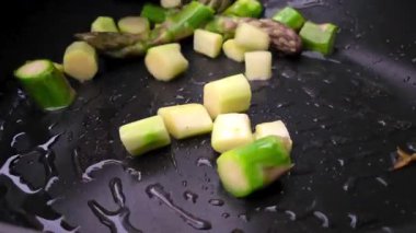 Tavada sotelenen yeni kesilmiş kuşkonmaz mızraklarının yakın çekimi. Canlı yeşil ve beyaz parçalar eşit dağılmış, yemek pişirme sürecinden kalan nem ile parıldıyor, taze ve sağlıklı bir mutfak temasını vurguluyor.