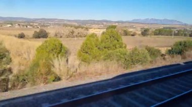 Trenin penceresinden çekilen sakin bir kırsal manzara, açık mavi gökyüzünün altında yemyeşil tarlalar, uzak dağlar ve canlı ağaçlar sergiliyor. Demiryolu rayları, seyahat ve keşifleri ima eden dinamik bir element sağlar..