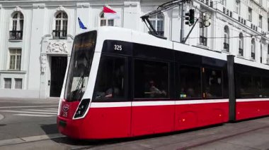 Viyana, Avusturya, 3 Mayıs 2025: Pürüzsüz bir kırmızı tramvay, tarihi mimariyle çelişen canlı bir Avrupa şehrinde seyahat ediyor. Yukarıda bayraklar dalgalanıyor, kültürel gurur. Yolcular, günlük şehir yaşamını ve toplu taşımayı gösteren içeriden görülebilir.