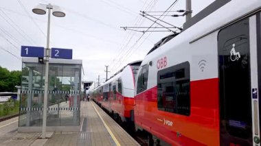 Vienna, Austria, May 3, 2025: A contemporary train halts at a european railway station, featuring distinct red, white, and black coloring. visible elements include the platform number sign, overhead wires, and surrounding greenery, suggesting