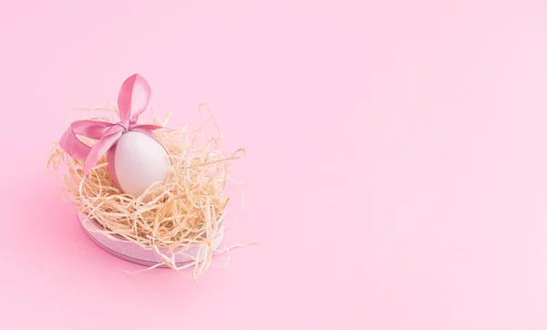 広いピンクイースターの背景 かわいい弓を持つ白い卵は わらの巣に横たわって 卵を寝ています ミニマルな春の休日のデザイン ストックフォト