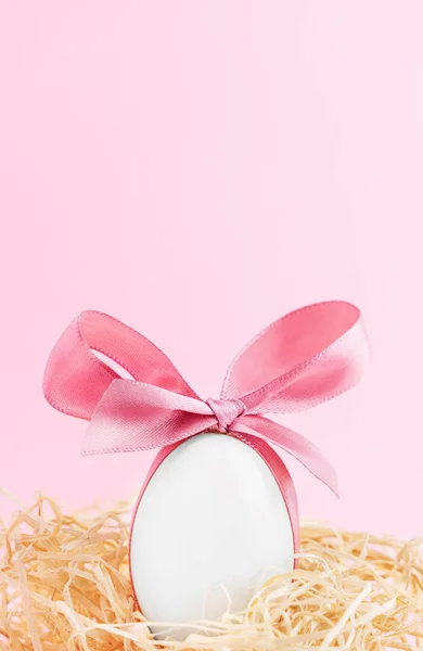 イースター休暇の背景 かわいいピンクの弓が鳥のわらの巣に座っている白い卵のクローズアップ ロイヤリティフリーのストック画像